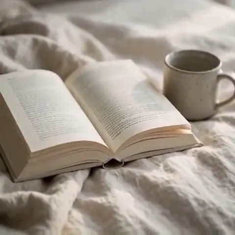 Pages Turn: Open hardback on crisp linen bedding beside a stoneware mug